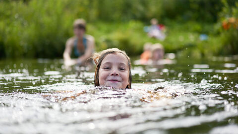 In Hessen gibt es viele Badeseen. Kind schwimmt glücklich im See. Im Hintergrund kommt auch die Familie ins Wasser.